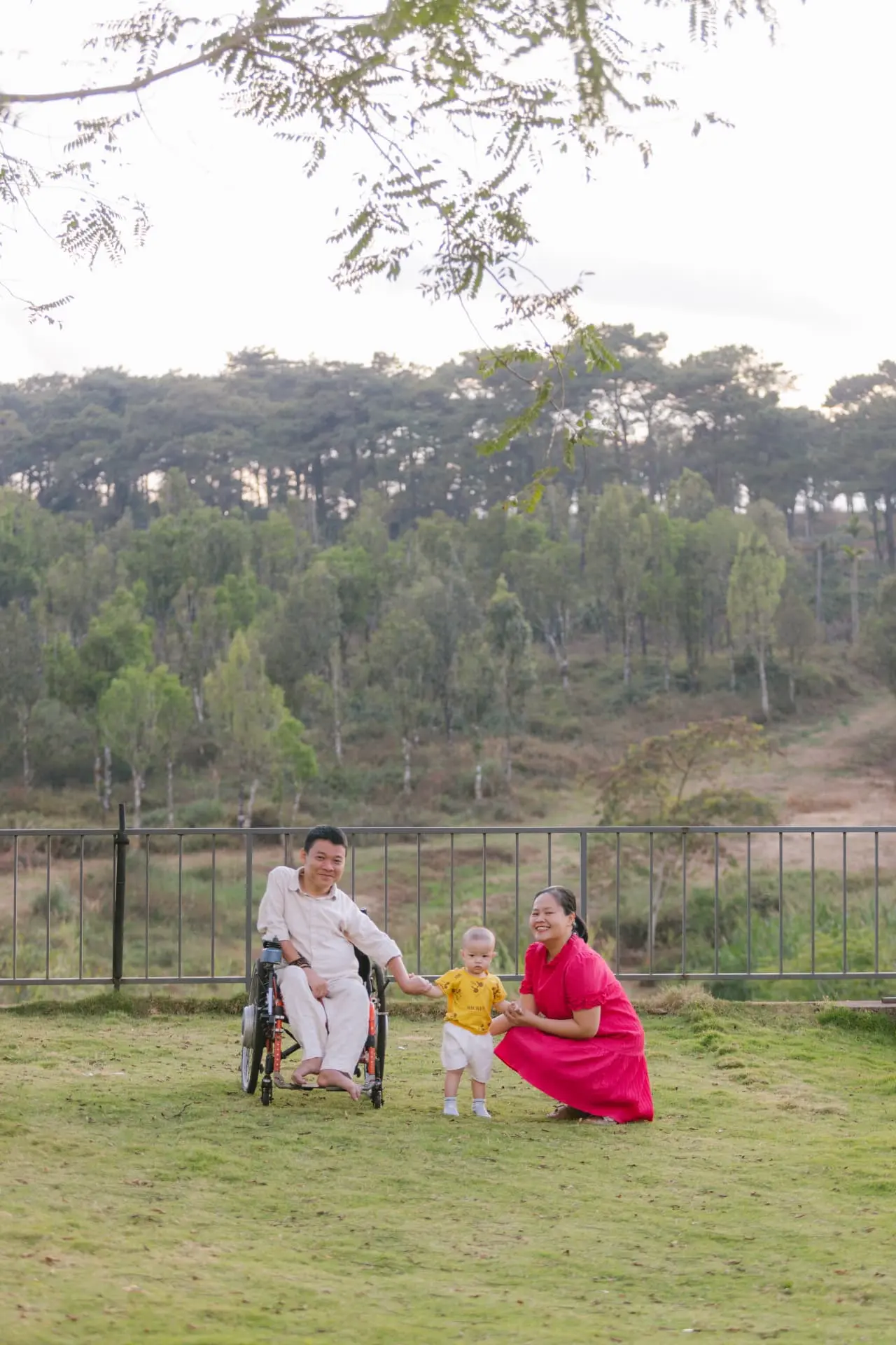 The three of them beneath a leafy branch in late afternoon light: father in his wheelchair holding his son's hand, mother kneeling beside the boy on a wide grass field