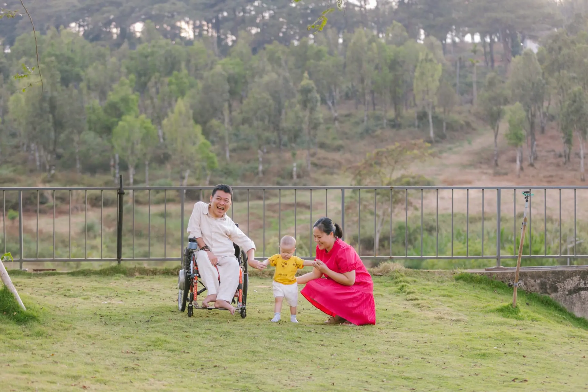 A father in a wheelchair laughing on a green lawn while his wife, kneeling in a red dress, holds their little son's hands as he learns to stand, with a pine hillside behind them