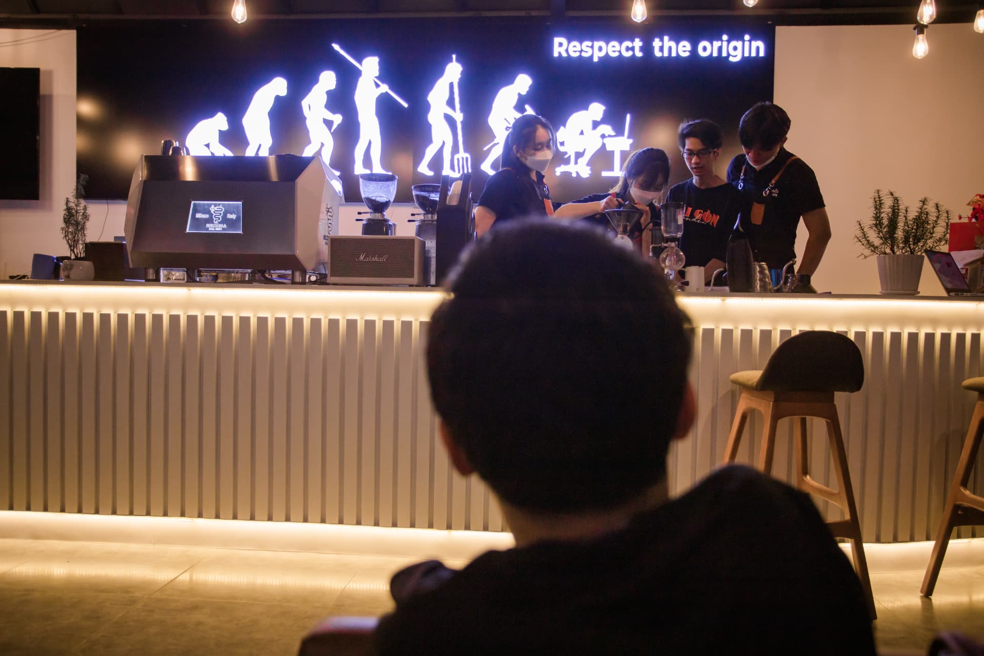 A quiet evening at the coffee bar, baristas working behind the counter under a lit ‘Respect the origin’ sign